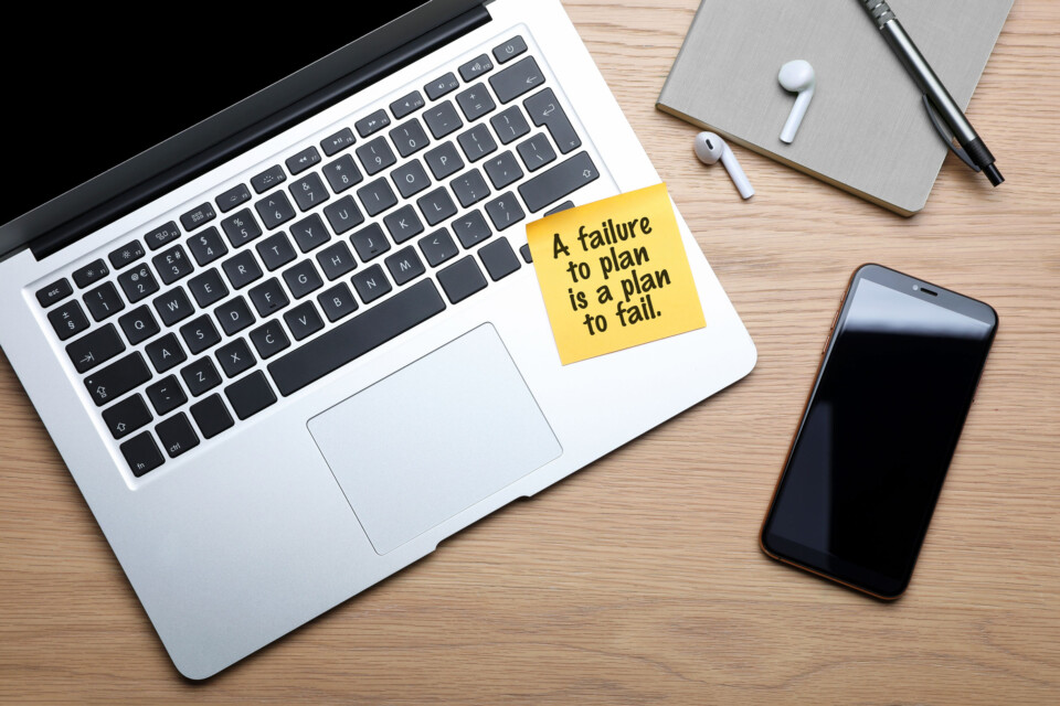 A wooden desk with a MacBook, iPhone, AirPods, notebook, pen, and a Post-it® note that reads, 